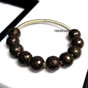 Könnte beinhalten: Ein Armband mit facettierten, dunkelrotbraunen Edelsteinperlen auf einem silberfarbenen Draht. Die Perlen haben ein gesprenkeltes Aussehen und sind gleichmäßig verteilt. Der Text "THEBEADSTONE" ist sichtbar. Ein schönes Schmuckstück.