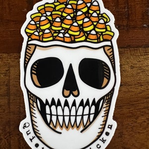 May include: A white skull sticker with a black outline and a toothy grin. The skull is filled with yellow and orange candy corn and the text "Quit at down chicken" is written around the bottom.