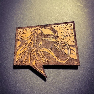 May include: A brown leather patch with a speech bubble shape. The patch features a detailed, engraved image of Venom, a Marvel Comics character, with a menacing expression. The background is textured with a speckled pattern.