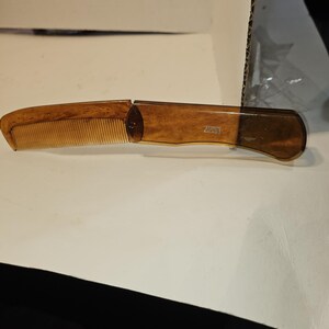 May include: A folding comb with a light brown, wood-grain appearance. The comb teeth are a lighter shade of brown. The handle is a darker brown and has the text "20031" printed on it. The comb is open and ready for use.