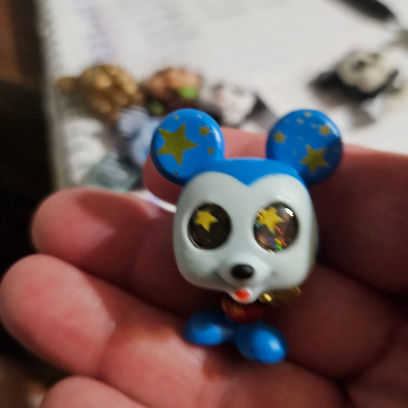 Peut inclure: Une petite figurine bleue et blanche de Mickey Mouse avec des &eacute;toiles dor&eacute;es sur ses oreilles et des yeux scintillants. Il porte une tenue rouge et bleue.