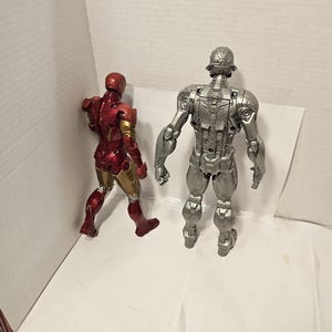 May include: Two action figures: Iron Man in red and gold, and Ultron in silver. The Iron Man figure is in a walking pose, while Ultron stands upright. Both figures are detailed and appear to be made of plastic.