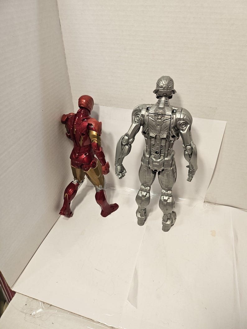 May include: Two action figures: Iron Man in red and gold, and Ultron in silver. The Iron Man figure is in a walking pose, while Ultron stands upright. Both figures are detailed and appear to be made of plastic.