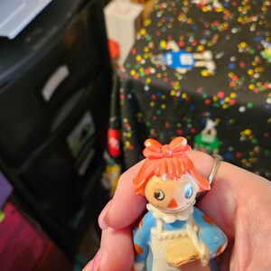 May include: A plastic Raggedy Ann doll with red hair, a blue dress, and a white apron. The doll is holding a small, round, yellow object in her hand.