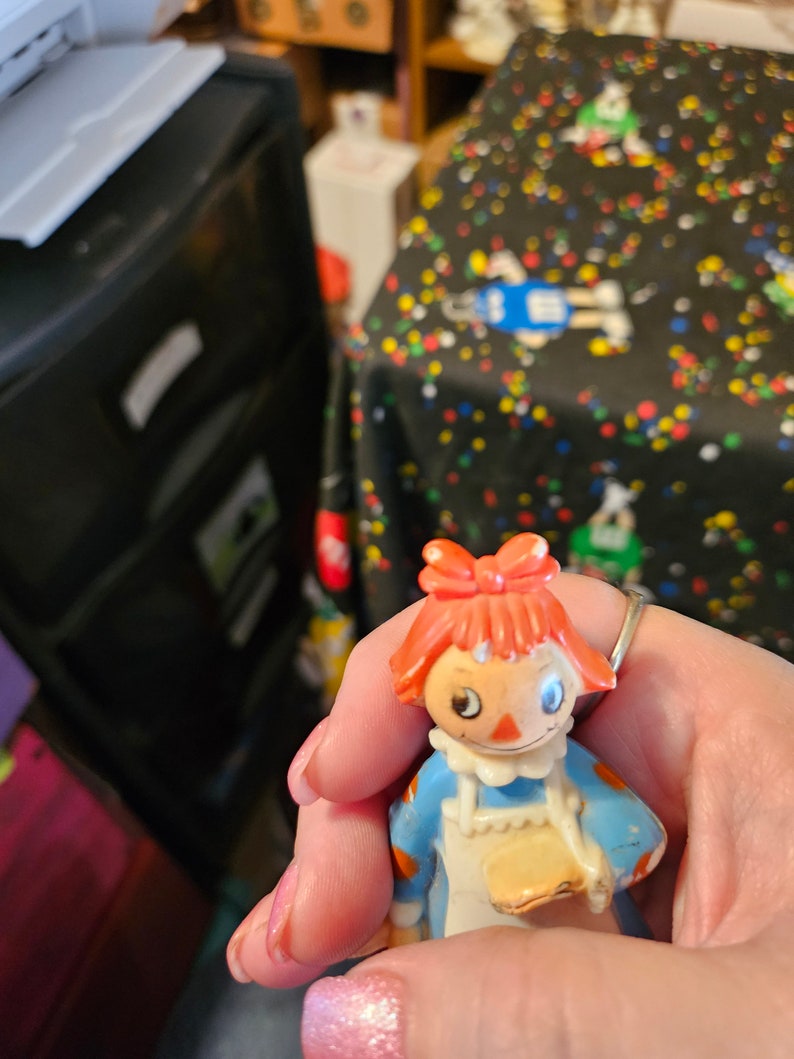 May include: A plastic Raggedy Ann doll with red hair, a blue dress, and a white apron. The doll is holding a small, round, yellow object in her hand.