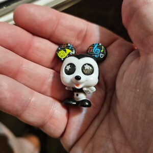 Peut inclure: Une petite figurine de Mickey Mouse noire et blanche avec des notes de musique et une trompette sur les oreilles. Il a de grands yeux brillants avec des &eacute;toiles blanches.