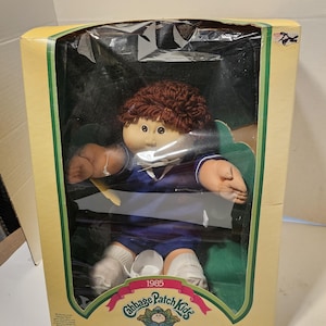 Könnte beinhalten: Eine Vintage Cabbage Patch Kids Puppe in ihrer Originalverpackung. Die Puppe hat braune Haare, ein blau-weißes Outfit und weiße Socken. Die Box ist gelb und grün mit dem Cabbage Patch Kids Logo und dem Text "1985". Für Kinder ab 3 Jahren.