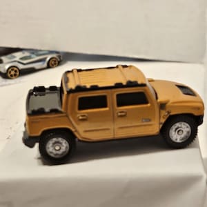 May include: A tan toy Hummer H2 SUV with black accents, featuring a detailed roof rack and realistic wheels. Another toy car is visible in the background. The toy car is a silver and blue sports car.