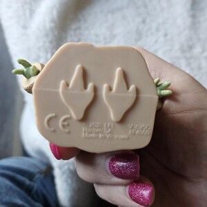 Puede incluir: Figura de color beige de un personaje con manos y orejas verdes. La parte inferior de la figura tiene dos huellas en forma de pie y el texto "CE", "&copy; 2022 LFL Hasbro SA Made In Vietnam".