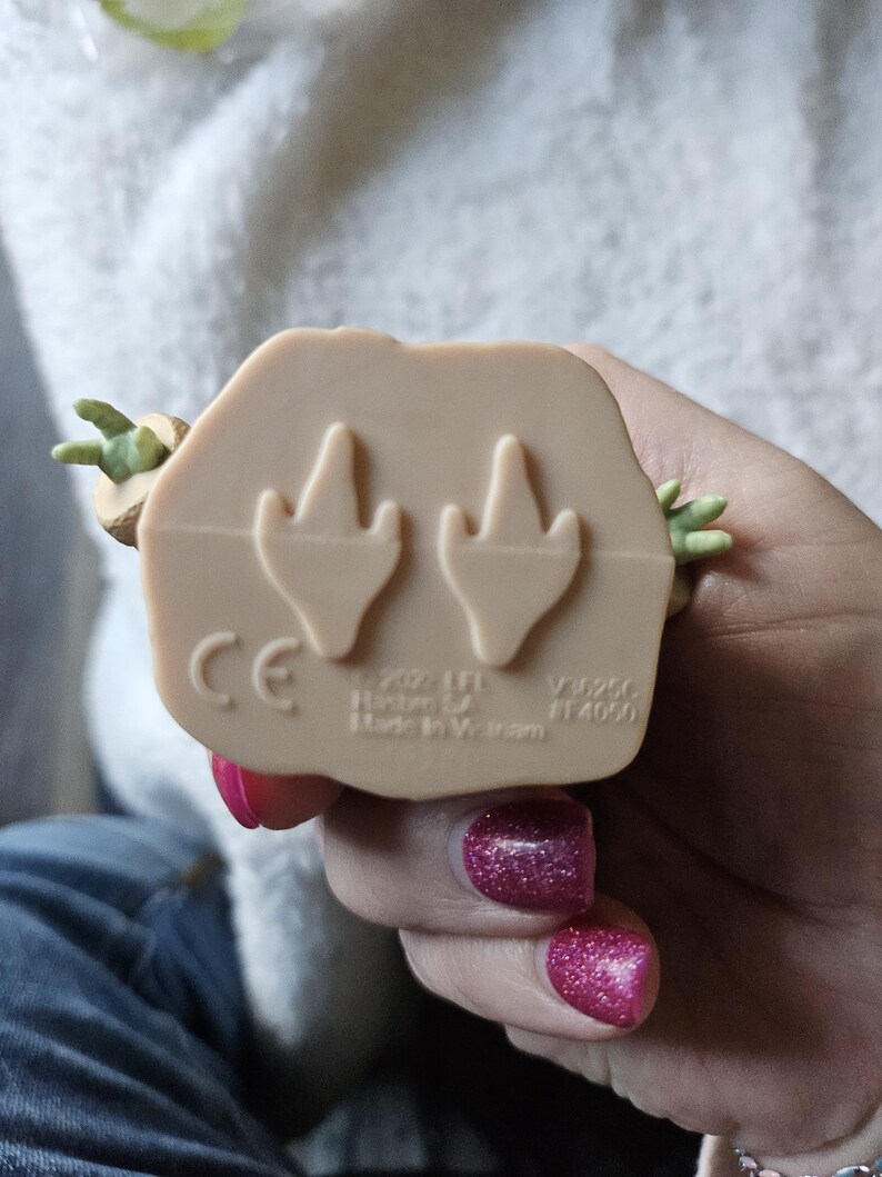Puede incluir: Figura de color beige de un personaje con manos y orejas verdes. La parte inferior de la figura tiene dos huellas en forma de pie y el texto "CE", "&copy; 2022 LFL Hasbro SA Made In Vietnam".