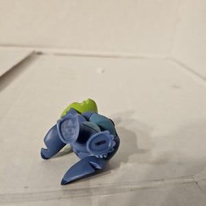 May include: A blue and green figurine of a cartoon character, lying on its back. The figurine has large ears, a small nose, and a wide mouth. The figurine is made of plastic and has a glossy finish. The figurine is a collectible item.