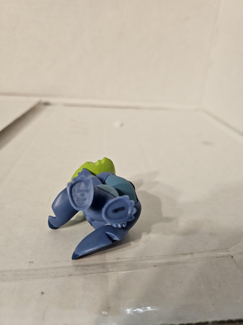 May include: A blue and green figurine of a cartoon character, lying on its back. The figurine has large ears, a small nose, and a wide mouth. The figurine is made of plastic and has a glossy finish. The figurine is a collectible item.