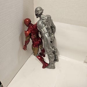 May include: Two action figures: Iron Man in red and gold armour, and Ultron in silver armour. The figures are posed together, with Iron Man leaning against a white surface. The figures are detailed and appear to be from a comic book or movie.