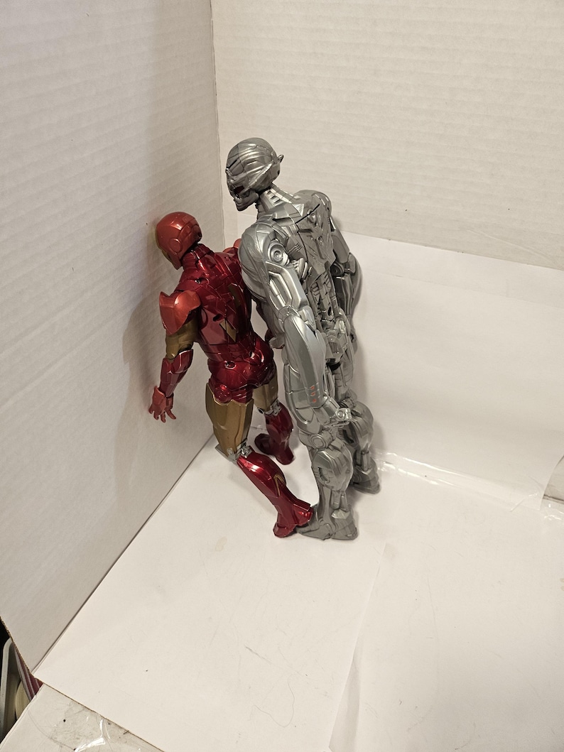 May include: Two action figures: Iron Man in red and gold armour, and Ultron in silver armour. The figures are posed together, with Iron Man leaning against a white surface. The figures are detailed and appear to be from a comic book or movie.