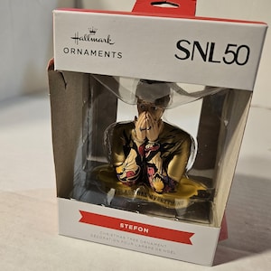 May include: Hallmark Christmas tree ornament featuring Stefon from SNL. The ornament is in a clear plastic case. Stefon is depicted in a gold outfit with a colorful design. The text "PLACE HAS MYBRYTHING" is visible.