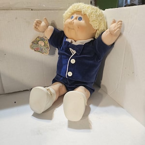 Può includere: Una bambola Cabbage Patch Kids con capelli biondi di filo, occhi blu e incarnato rosato. La bambola indossa un completo di velluto blu scuro con bottoni e scarpe bianche. Un'etichetta con il logo Cabbage Patch Kids è attaccata.