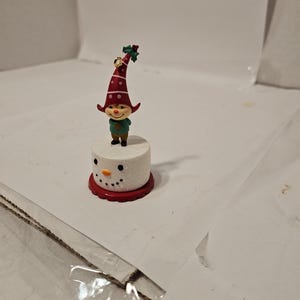 May include: A whimsical Christmas figurine featuring a snowman base with a red base and a smiling elf wearing a red hat with white polka dots. The elf has a green shirt and black shoes. The hat is adorned with holly.