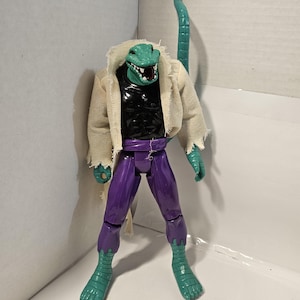May include: A detailed action figure of a reptilian humanoid character. The figure features a green reptilian head with sharp teeth, a black torso, purple pants, and green reptilian feet. It wears a tattered, off-white jacket and has a long, green tail.