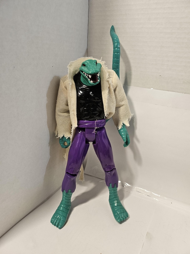 May include: A detailed action figure of a reptilian humanoid character. The figure features a green reptilian head with sharp teeth, a black torso, purple pants, and green reptilian feet. It wears a tattered, off-white jacket and has a long, green tail.