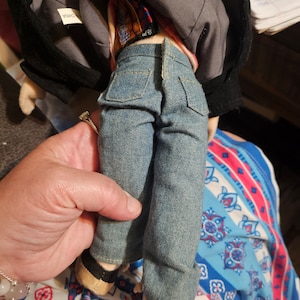 K&ouml;nnte beinhalten: Eine Pl&uuml;schpuppe, die eine blaue Jeansjacke, ein rot-wei&szlig; gestreiftes Hemd und eine blaue Jeans tr&auml;gt. Die Puppe h&auml;lt einen kleinen, braunen Pl&uuml;schhund in ihren Armen. Die Puppe ist aus weichem, pl&uuml;schem Stoff gefertigt und hat einen freundlichen Gesichtsausdruck.