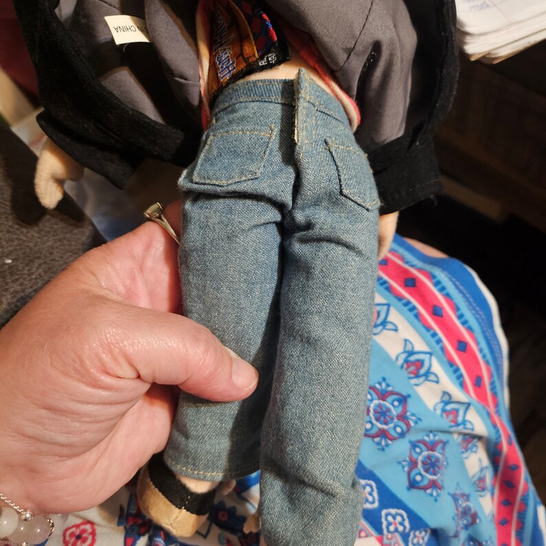 K&ouml;nnte beinhalten: Eine Pl&uuml;schpuppe, die eine blaue Jeansjacke, ein rot-wei&szlig; gestreiftes Hemd und eine blaue Jeans tr&auml;gt. Die Puppe h&auml;lt einen kleinen, braunen Pl&uuml;schhund in ihren Armen. Die Puppe ist aus weichem, pl&uuml;schem Stoff gefertigt und hat einen freundlichen Gesichtsausdruck.