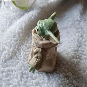 Puede incluir: Una figura de piel verde y orejas grandes con una t&uacute;nica color beige. La figura tiene ojos grandes y est&aacute; posando con un brazo extendido. La figura es un personaje de una popular franquicia de ciencia ficci&oacute;n. La figura est&aacute; sentada sobre una superficie blanca y esponjosa.