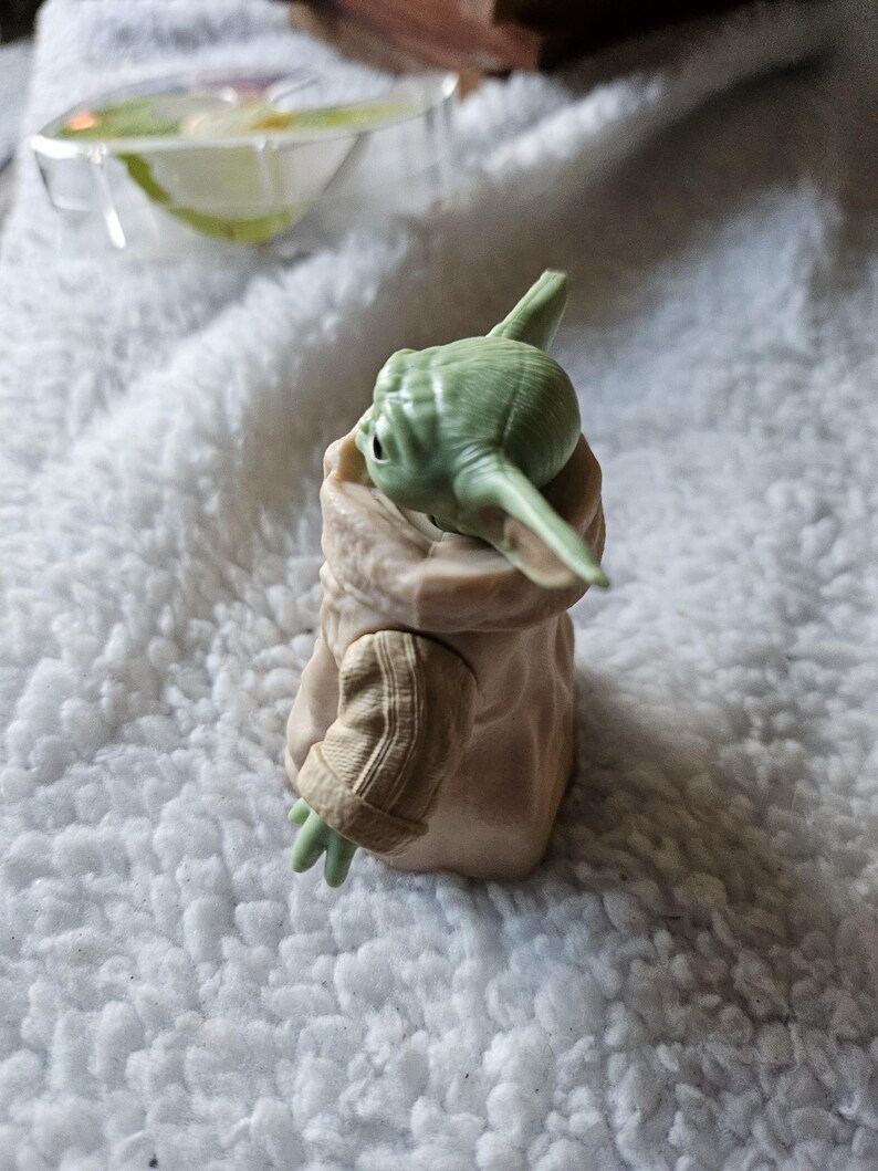 Puede incluir: Una figura de piel verde y orejas grandes con una t&uacute;nica color beige. La figura tiene ojos grandes y est&aacute; posando con un brazo extendido. La figura es un personaje de una popular franquicia de ciencia ficci&oacute;n. La figura est&aacute; sentada sobre una superficie blanca y esponjosa.