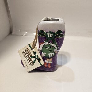 May include: A purple and white M&M's candy boot-shaped ornament with a green M&M's character wearing a Santa hat. The ornament has a tag that reads "M&M's Milk Chocolate Chocolate Candy" and weighs 1.0 oz (28.4g).