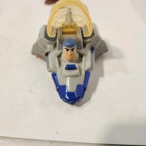 May include: A toy spaceship with a clear yellow canopy and a blue and gray body. Inside the spaceship is a small Buzz Lightyear figure with a blue helmet. The toy is on a white surface.