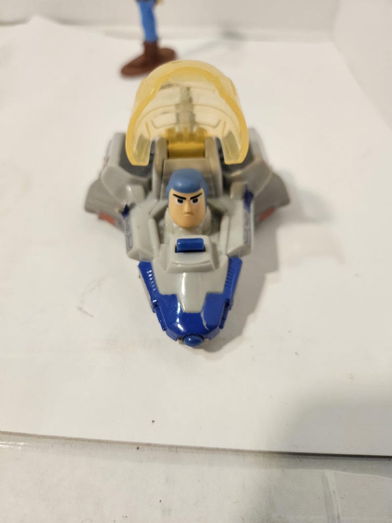 May include: A toy spaceship with a clear yellow canopy and a blue and gray body. Inside the spaceship is a small Buzz Lightyear figure with a blue helmet. The toy is on a white surface.