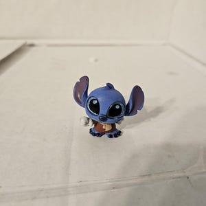 Pu&ograve; includere: Una piccola statuina blu del personaggio Stitch del franchise Lilo & Stitch. La statuina ha grandi occhi espressivi con accenti glitterati, grandi orecchie e un abito marrone. La statuina &egrave; su una superficie bianca.