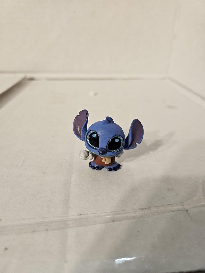Pu&ograve; includere: Una piccola statuina blu del personaggio Stitch del franchise Lilo & Stitch. La statuina ha grandi occhi espressivi con accenti glitterati, grandi orecchie e un abito marrone. La statuina &egrave; su una superficie bianca.