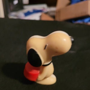 May include: A beige plastic figurine of Snoopy holding a red heart-shaped object. The figurine is a popular character from the Peanuts comic strip.