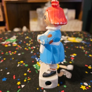 May include: A vintage plastic doll pencil sharpener with a blue dress, white apron, and red hair. The doll is standing on a white base with a pencil sharpening hole.