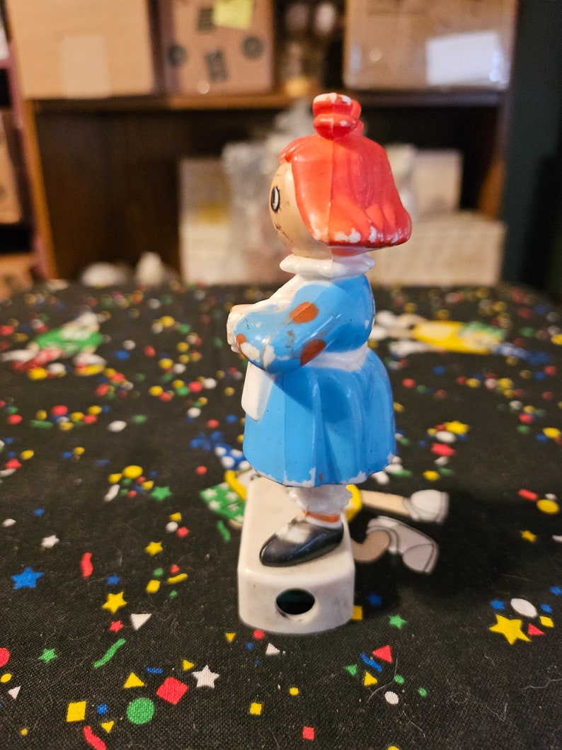 May include: A vintage plastic doll pencil sharpener with a blue dress, white apron, and red hair. The doll is standing on a white base with a pencil sharpening hole.