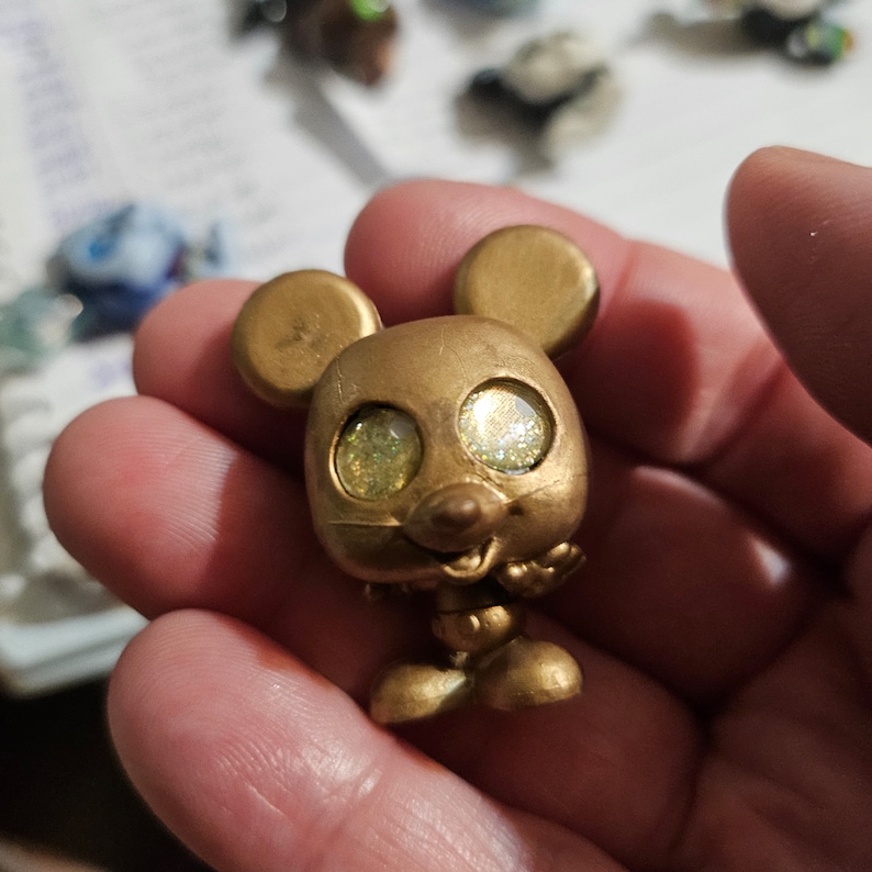 Peut inclure: Une figurine en plastique dor&eacute;e de Mickey Mouse avec des yeux verts scintillants. La figurine l&egrave;ve les mains en signe de salut.