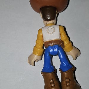 Op de afbeelding: Een speelgoed cowboyfiguur, van achteren gezien. Het draagt een bruine hoed, geel overhemd, wit vest, blauwe broek en bruine laarzen. Dit is een kinderspeelgoed.