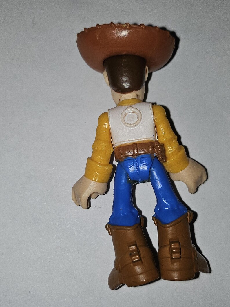 Op de afbeelding: Een speelgoed cowboyfiguur, van achteren gezien. Het draagt een bruine hoed, geel overhemd, wit vest, blauwe broek en bruine laarzen. Dit is een kinderspeelgoed.