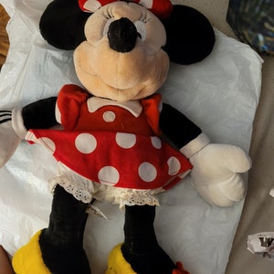 Puede incluir: Un juguete de peluche de Minnie Mouse con un vestido rojo con lunares blancos y zapatos amarillos con lazos rojos.