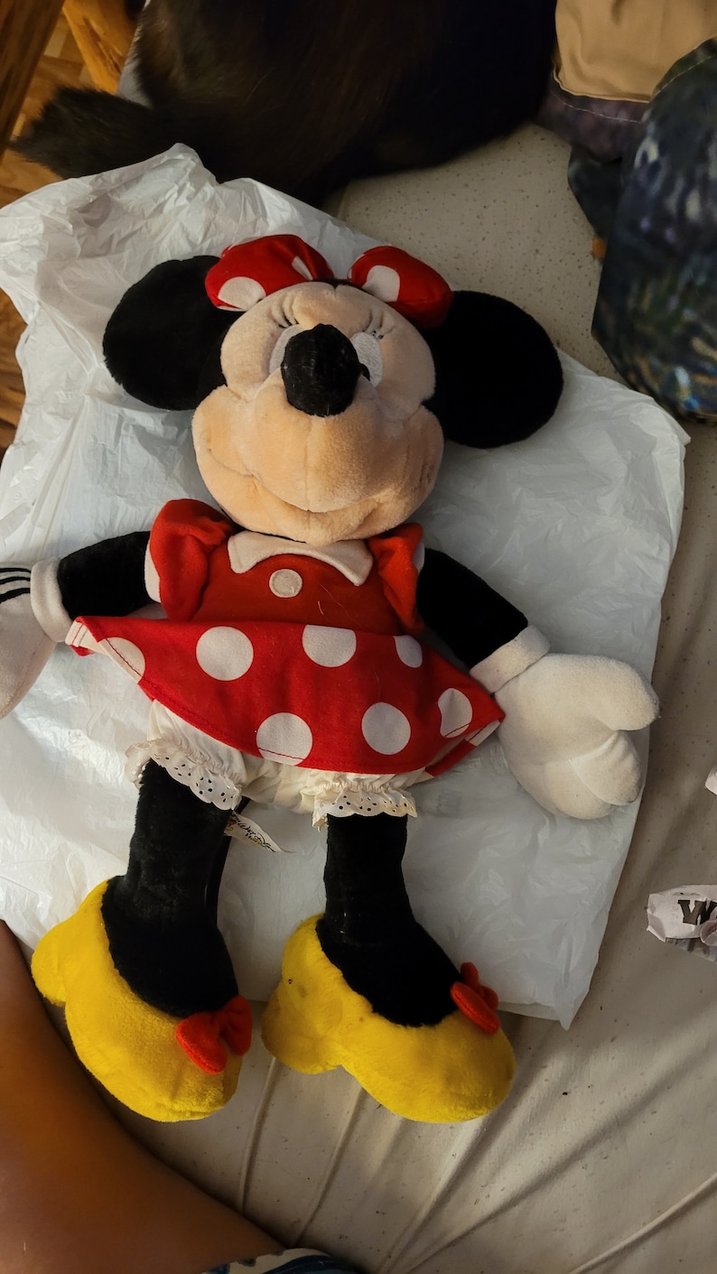 Puede incluir: Un juguete de peluche de Minnie Mouse con un vestido rojo con lunares blancos y zapatos amarillos con lazos rojos.