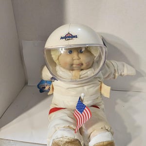 May include: A vintage Cabbage Patch Kids Astronaut doll, wearing a white spacesuit and helmet with the word "Astronauts" printed on the helmet. The doll is holding a small American flag. The doll has blonde hair and a tag attached to its left arm.