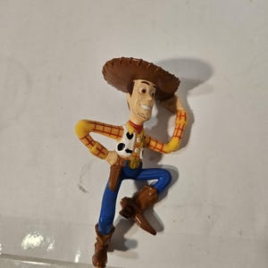 May include: A plastic figurine of Woody from Toy Story. The figure is wearing a brown cowboy hat, a yellow and red plaid shirt, a white vest with a black cow print, blue pants, and brown boots. The figure is in a dynamic pose.