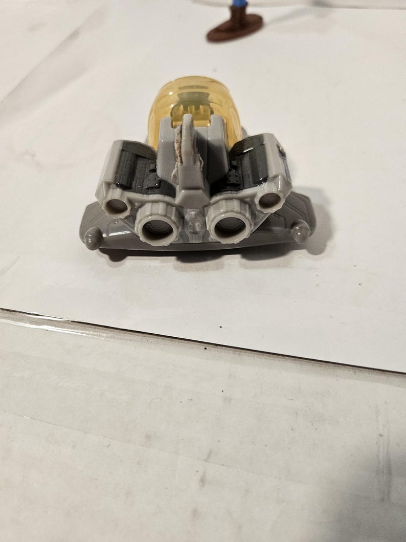 May include: A gray and yellow toy spaceship with multiple exhaust ports and a clear, yellow dome. The toy has a detailed design with various mechanical components, suggesting a futuristic or space-themed toy.