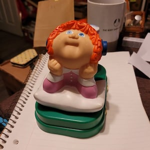 May include: A plastic Cabbage Patch Kid doll wearing a pink dress and blue headphones, sitting on a white pillow on a green base.