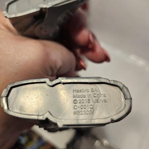 May include: Two silver toy figures. The base of one figure has the text "Hasbro SA Made in China &copy;2015 Marvel C-001C #B2303". The figures are held in a person's hand.