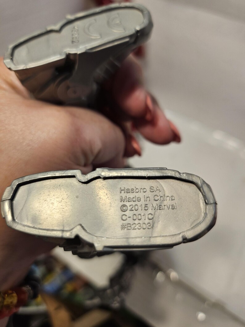 May include: Two silver toy figures. The base of one figure has the text "Hasbro SA Made in China &copy;2015 Marvel C-001C #B2303". The figures are held in a person's hand.