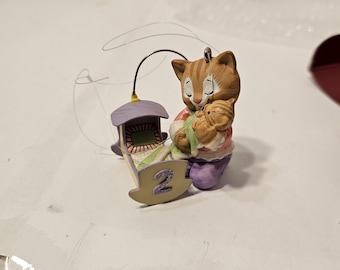 May include: A ceramic ornament featuring a mother cat holding a kitten, with a small rocking cradle. The ornament is painted in soft pastel colors, with the number "2" on the cradle. A wire loop is attached for hanging.