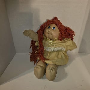 Op de afbeelding: Een vintage Cabbage Patch Kid pop met rood garen haar, blauwe ogen en een gele jurk met witte kant.