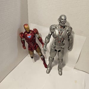 May include: Two action figures: Iron Man in red and gold armour, and Ultron in silver armour. Iron Man is posed with one leg forward, while Ultron stands upright. Both figures are on a white surface.