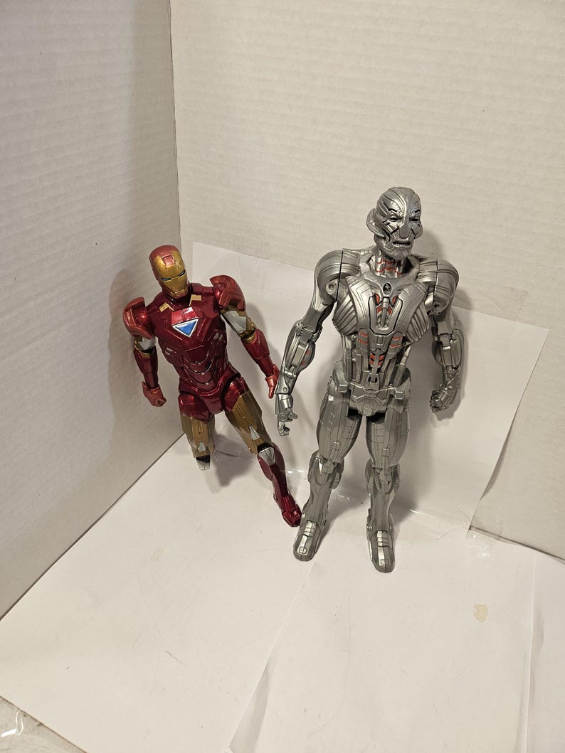 May include: Two action figures: Iron Man in red and gold armour, and Ultron in silver armour. Iron Man is posed with one leg forward, while Ultron stands upright. Both figures are on a white surface.